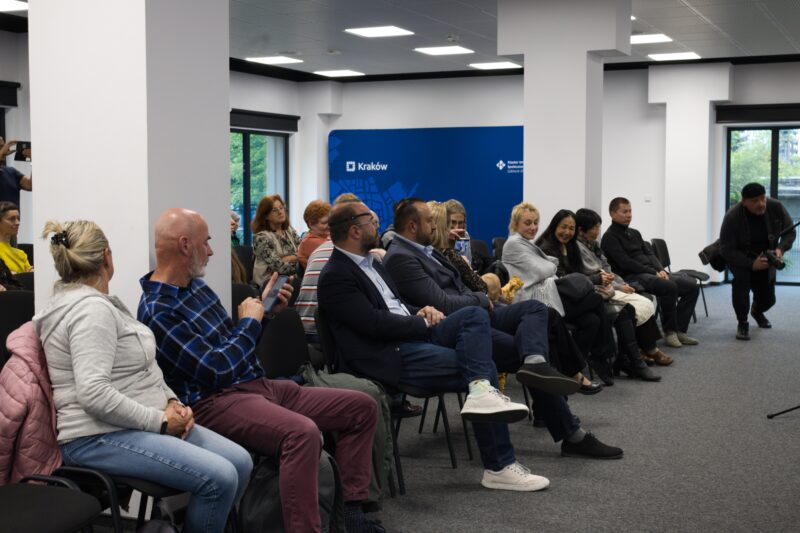 niewielka sala z białymi filarami, na pierwszym planie rząd krzeseł z publicznością