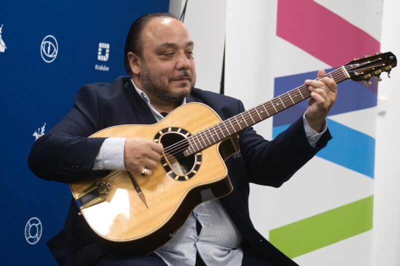 mężczyzna grający na gitarze, w tle fragment niebieskiego baneru z logotypami Krakowa i Małopolski
