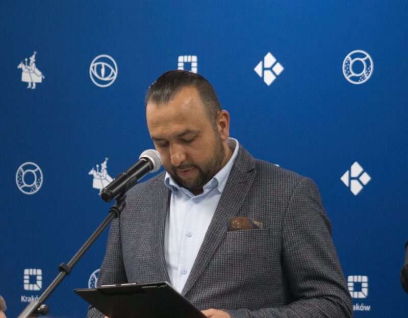 mężczyzna przy mikrofonie czytający tekst przemówienia, na niebieskim tle z logotypami Krakowa