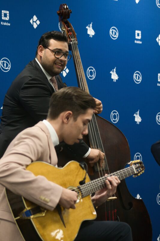 mężczyzna grający na kontrabasie, przed nim młody muzyk grający na gitarze, w tle niebieski baner z logotypami Krakowa