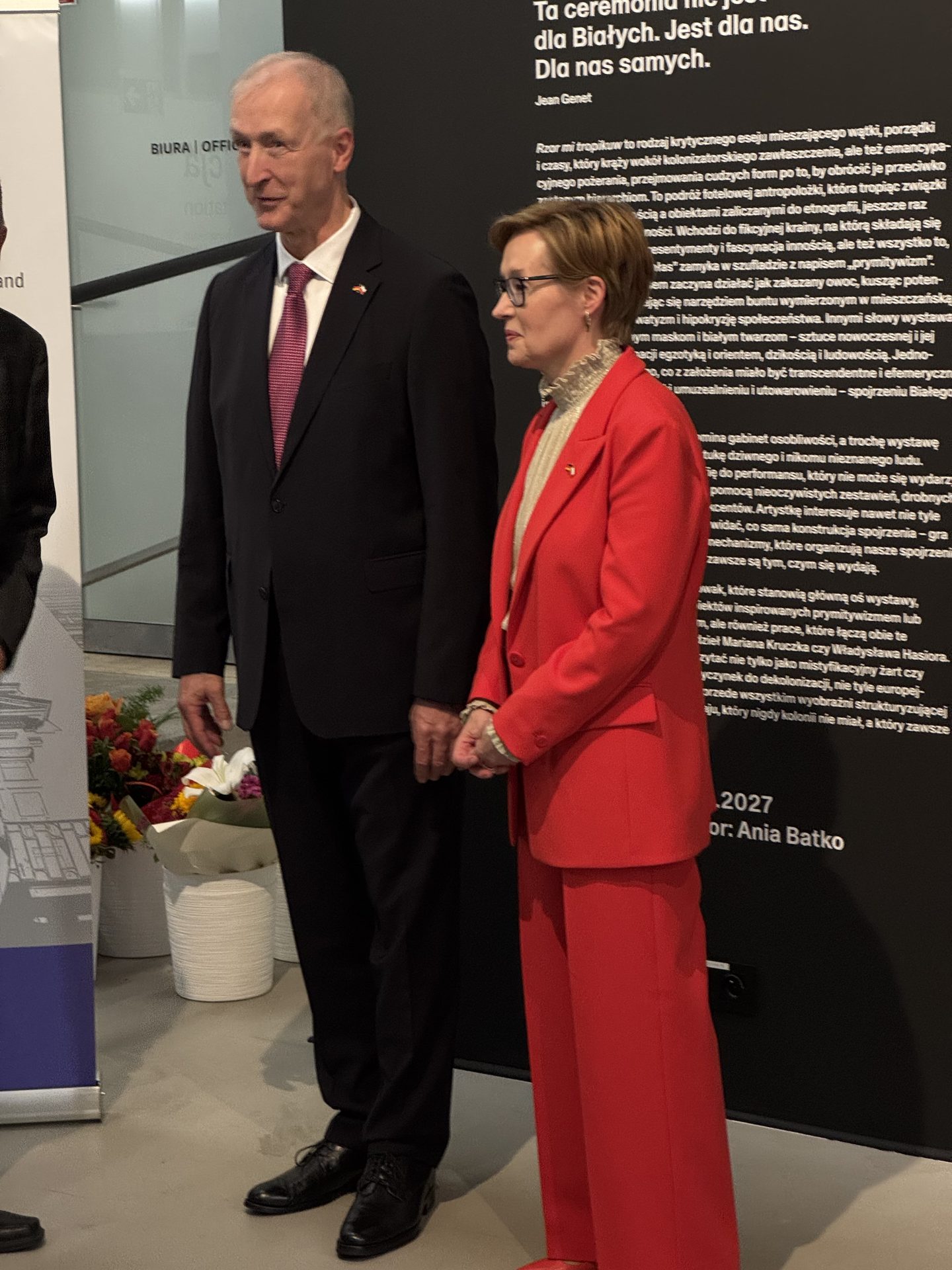 Dwójka elegancko ubranych osób stoi obok siebie podczas wydarzenia. Mężczyzna ma na sobie ciemny garnitur i różowy krawat, a kobieta — czerwony garnitur i okulary. W tle widać wystawę z polskim tekstem oraz donicę z kwiatami.